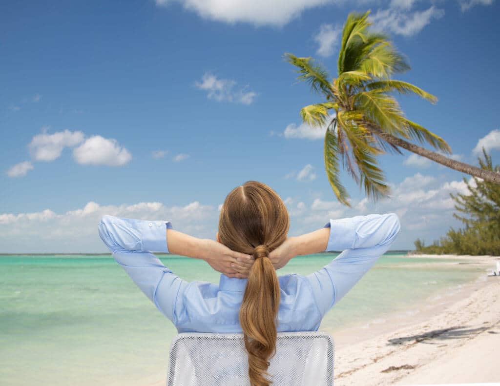 A woman reclining on a beach chair looking out at a tropical beach and ocean with palm tree in background