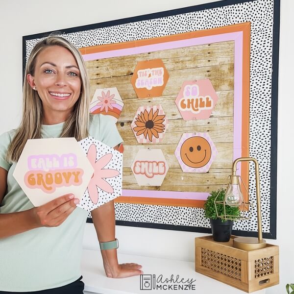 A teacher holding up two Retro Fall Classroom Decor posters in front of a decorated bulletin board featuring more of the hexagon shaped fall themed posters.