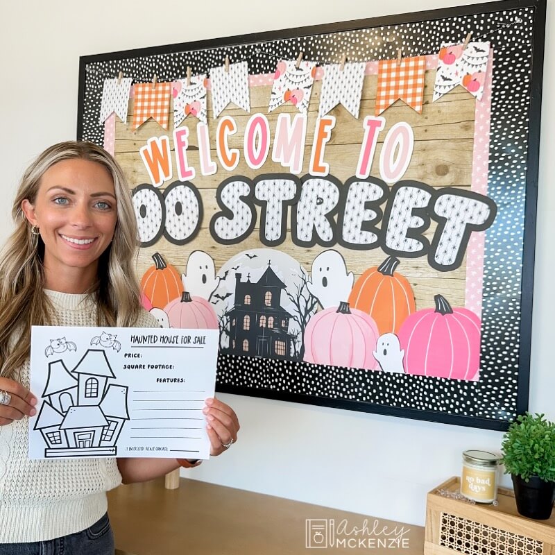 A teacher holding up a writing template that says "Haunted House for Sale" standing in front of a bulletin board decorated with the saying "Welcome to Boo Street" Halloween Classroom Decor