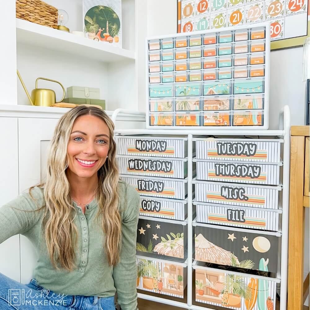 A teacher sitting in front of a teacher toolbox and a 12-drawer rolling cart - both are decorated with tropical themed organizing labels.