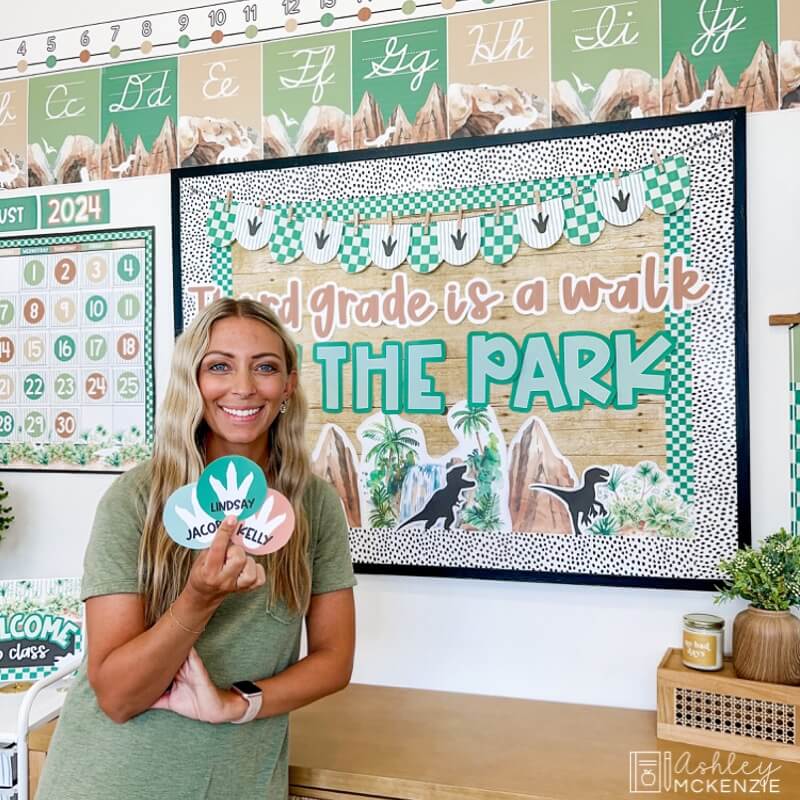 A teacher standing in front of a classroom bulletin board decorated with a dinosaur theme that says "Third grade is a walk in the park"