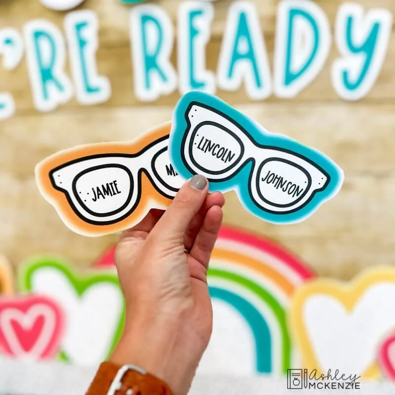 A teacher is holding up two cutouts of glasses for a bulletin board that each have a students' name printed on them