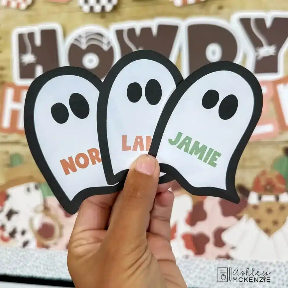 Three text editable ghost cutouts are held in front of a Halloween classroom decor bulletin board with students' names printed on them