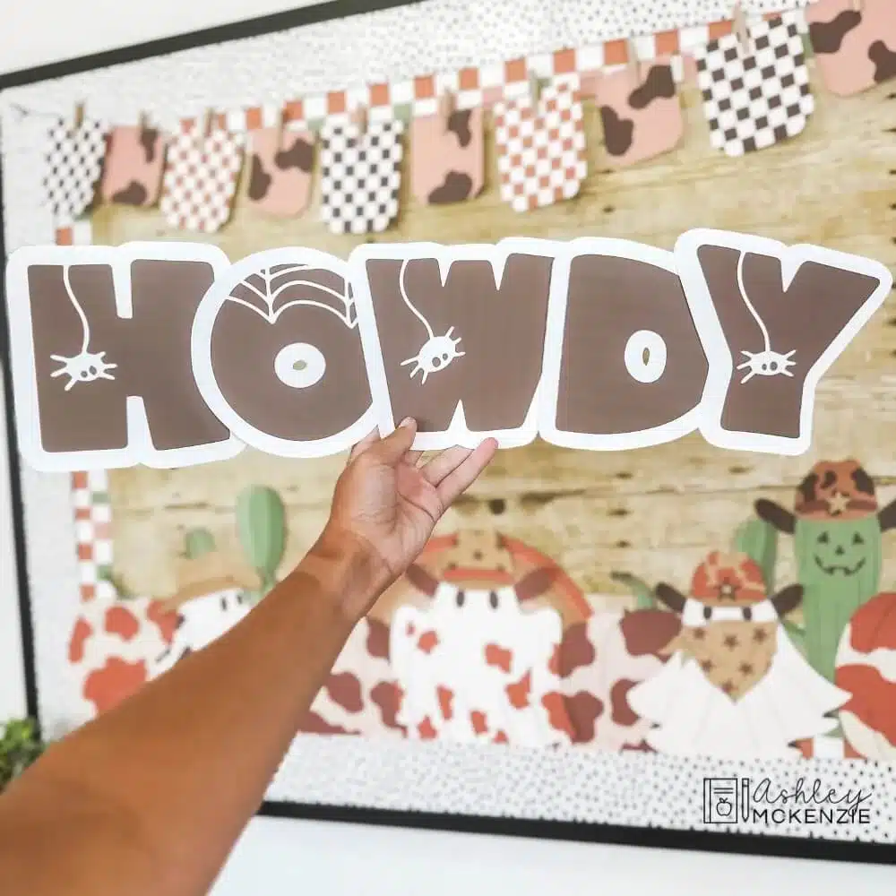 Large bulletin board letters in a spooky font spell the word "Howdy." They are held in front of a bulletin board decorated for Halloween.