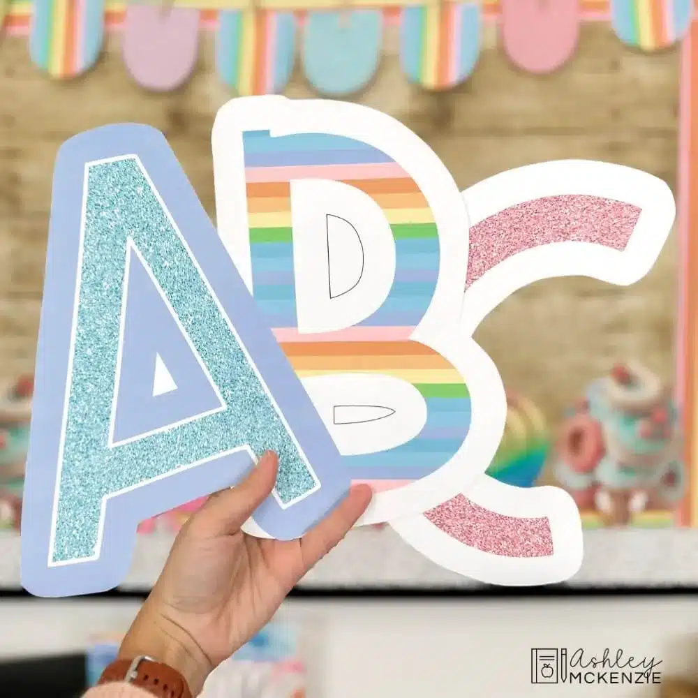 3 large bulletin board letters are being held up in front of a classroom bulletin board featuring glittery pastel designs