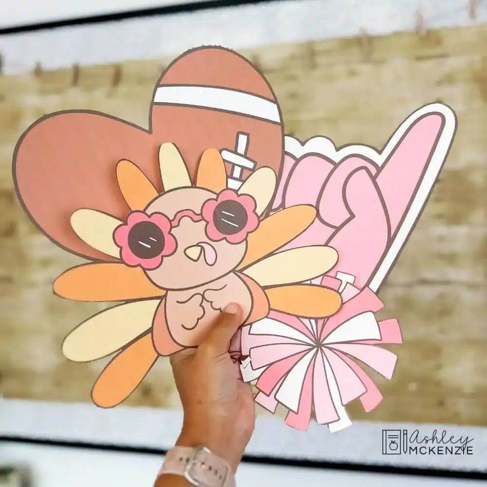 Bulletin board cutouts are held in front of a classroom display including a turkey wearing sunglasses, a heart shaped football, pompom, and foam finger