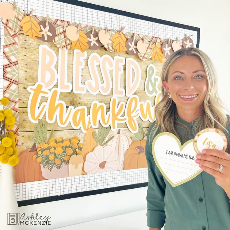 A teacher holding up a student "I am thankful for..." activity and standing in front of a classroom bulletin board decorated for Thanksgiving