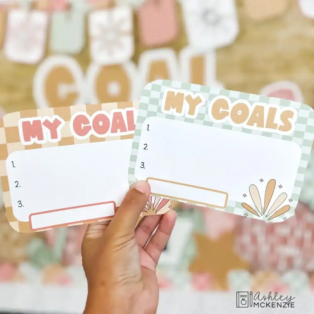 New Year Goals writing templates are held in front of a decorated classroom bulletin board; the heading "My Goals" is shown on each template with space to write student goals.