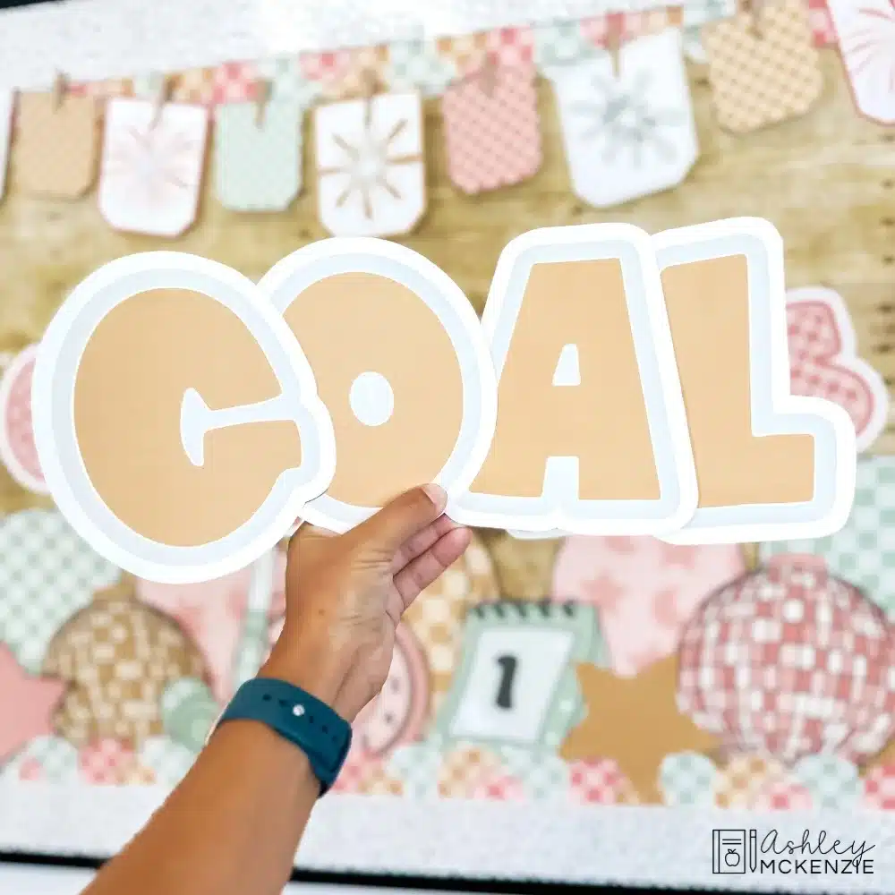 A hand is holding up the letters to spell "Goal" in front of a decorated classroom New Year bulletin board