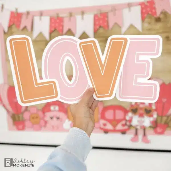 love-is-in-the-air-bb-bundle-Slide17 A hand holding up large bulletin board letter cutouts that spell "LOVE" in front of a Valentine's Day bulletin board display in a classroom