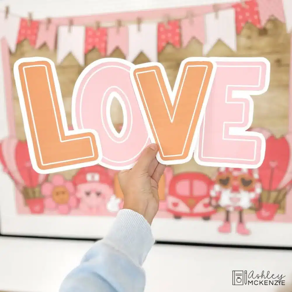 The letters for "LOVE" are held in front of a Valentine's Day bulletin board display in a classroom