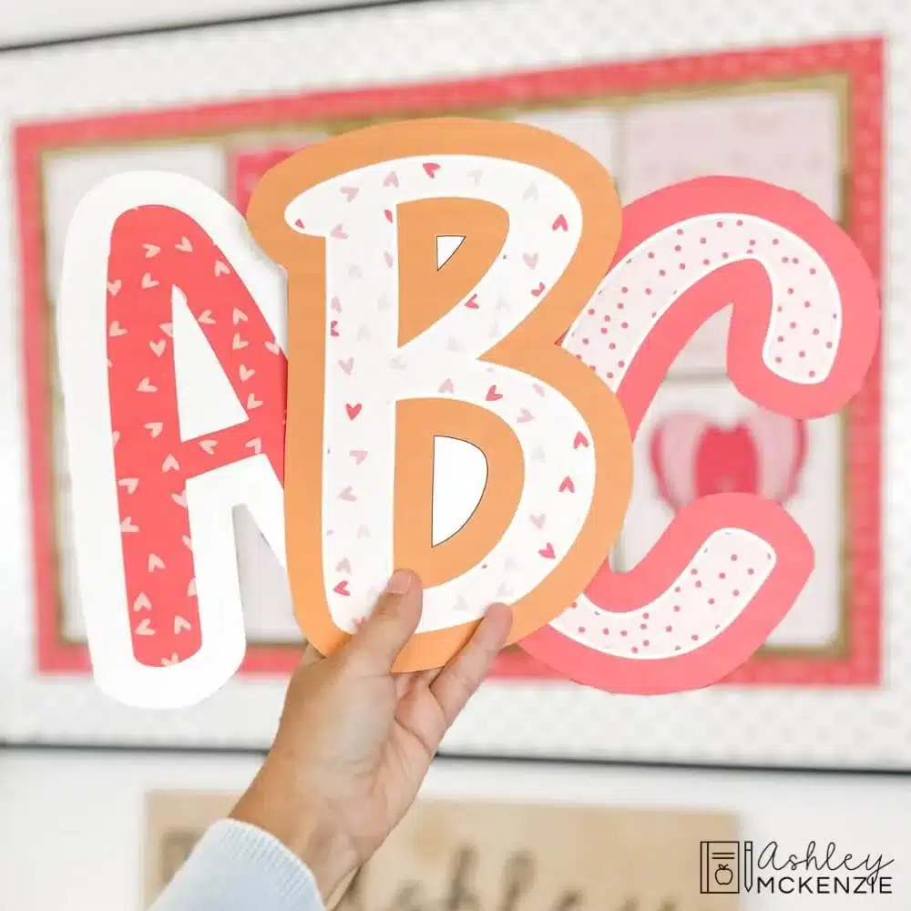 Large printable bulletin board letters A, B, and C are shown in front of a Valentine's Day bulletin board display; the letters feature heart patterns and classic Valentine colors