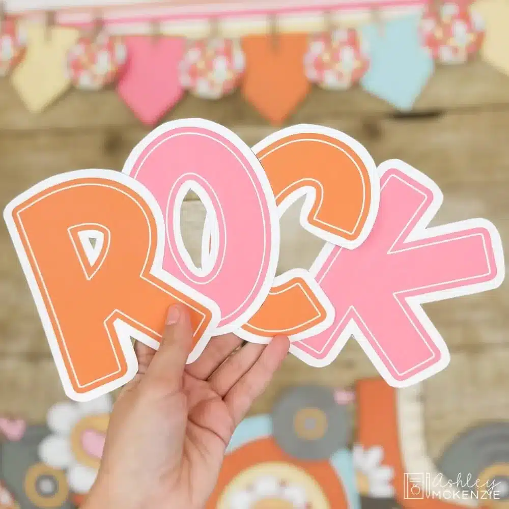 A hand holds letter cutouts that spell the word "ROCK" in bright orange and pink