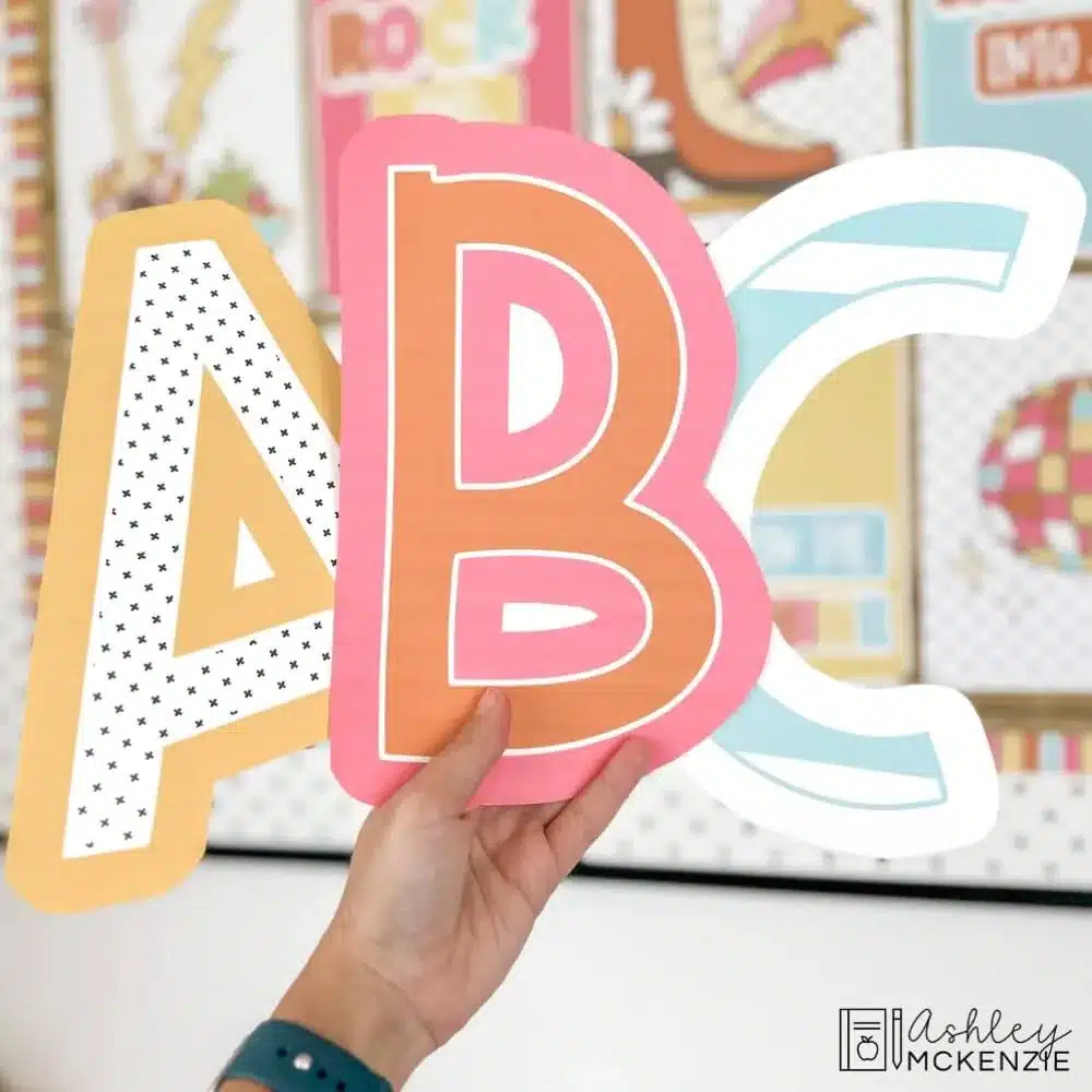 A hand holds 3 large bulletin board letter cutouts of A, B, and C in spring themed colors and prints