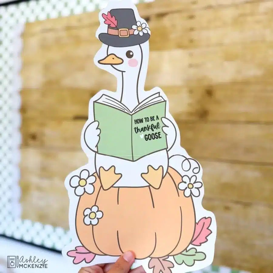 A Thanksgiving classroom decor cutout of a goose wearing a pilgrim hat sitting on top of a pumpkin reading a book