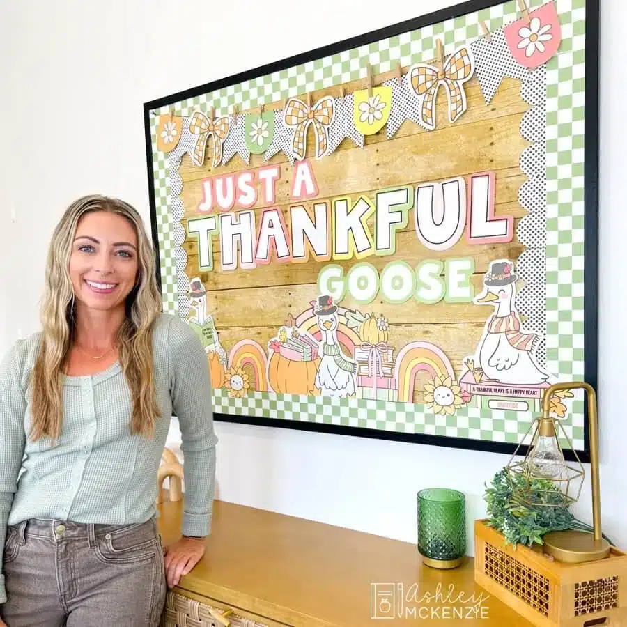 A teacher standing in front of a November bulletin board decorated with Just a Thankful Goose on it for Thanksgiving classroom decor