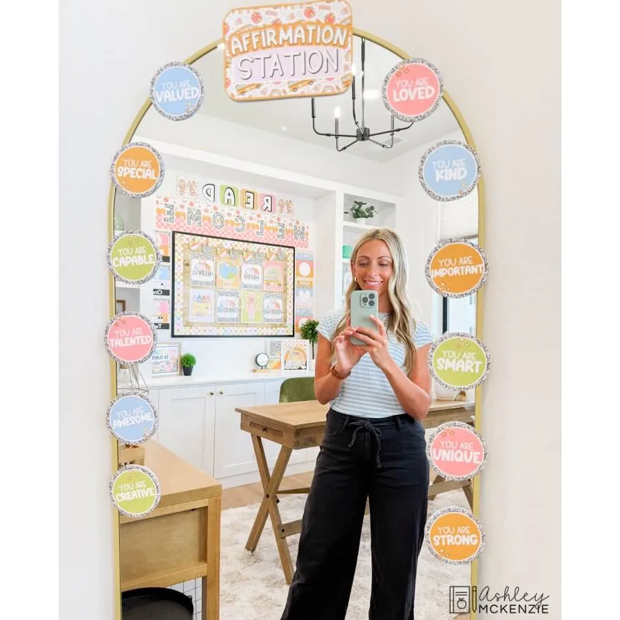 A teacher stands in front of a mirror decorated with a pastel classroom decor themed affirmation station kit